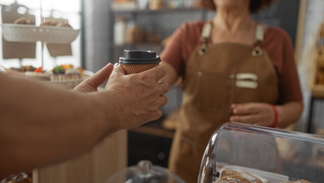 Woman in a bakery hands a cup of coffee to a man over the counter indoors, highlighting their professional interaction and cozy shop environment.