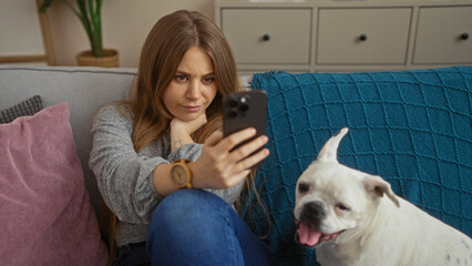 A young blonde caucasian woman sitting with her pet canine in a living room, using a smartphone at home.