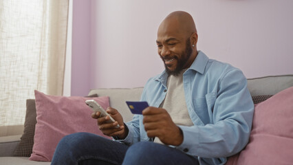 Handsome young african american man sitting in a living room smiling while using a phone and holding a credit card indoors