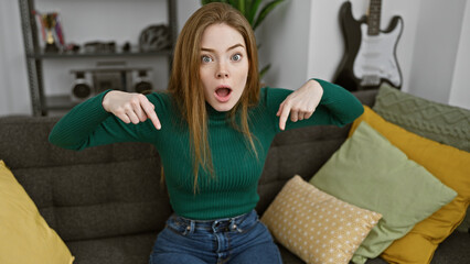 Surprised young woman in sweater pointing at herself indoors with guitar in background.