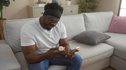 Handsome young african american man with a beard sits in a living room, examining a medication bottle while using a smartphone.