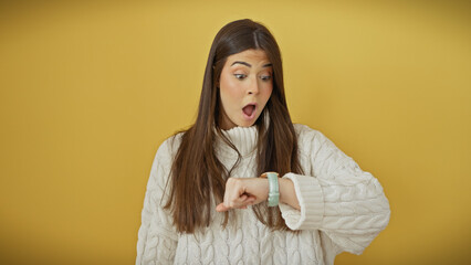 Surprised young woman checking the time on her wristwatch against a solid yellow background.