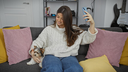 Smiling woman taking selfie with terrier dog on a cozy sofa in a modern living room.