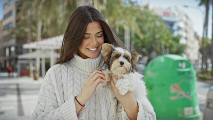 A smiling young hispanic woman holds a biewer yorkshire terrier on a sunlit urban street