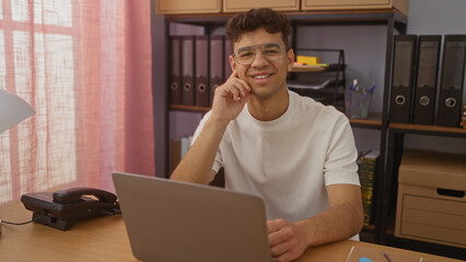 Handsome young hispanic man sitting at an office desk with a laptop and smiling confidently in a well-lit, organized workplace interior.