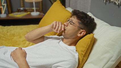 Young hispanic man lying on a bed in a bedroom wearing glasses and a white shirt, with a thoughtful expression and a hand on his mouth, indoors at home.