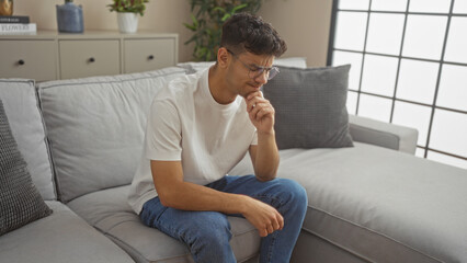 Young hispanic man in glasses sitting thoughtfully on a sofa in a modern living room, wearing a white t-shirt and blue jeans, indoors at home.