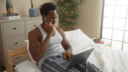 Young man talking on phone while using laptop in bedroom