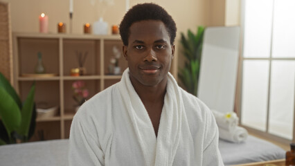 Handsome young man in a spa wellness room with candles in the background and wearing a white robe, sitting indoors and looking directly at the camera