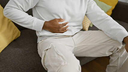 African american man in white clothing sitting indoors on a gray couch holding stomach, suggesting discomfort or pain.