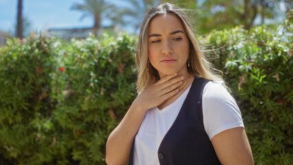 A beautiful young caucasian woman touching her throat while standing outdoors in a bright urban park during the day.