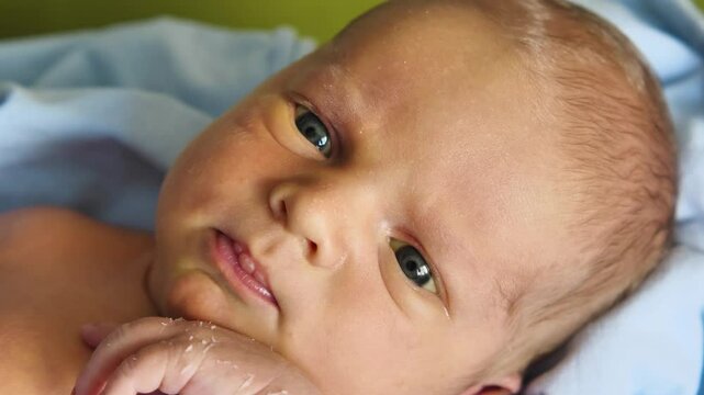 A close-up of a newborn baby boy's face. The infant has dark hair, dark eyes and pink lips with calluses from sucking on the breast. The skin on his arm is peeling