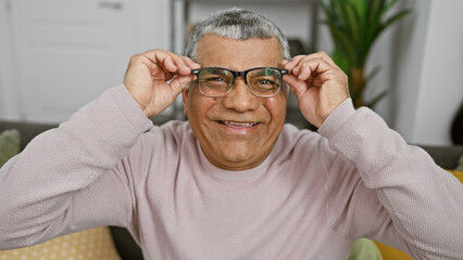 A smiling mature man adjusts his glasses in a cozy living room, exuding warmth and contentment. - Powered by Adobe