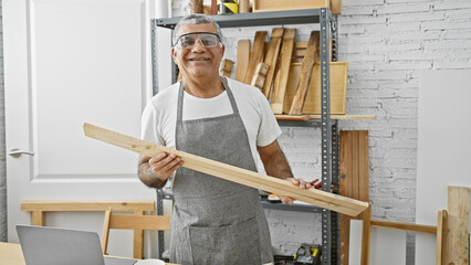 A smiling mature man in an apron holding lumber in a well-organized carpentry workshop.