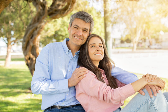 A man and woman relax together outdoors, sitting on the grass beneath a large tree as sunlight filters through the leaves, creating a warm and inviting atmosphere.