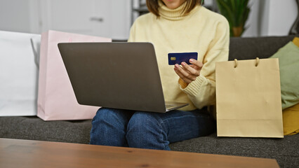 A woman sitting indoors with a laptop and credit card, possibly shopping online next to shopping bags.