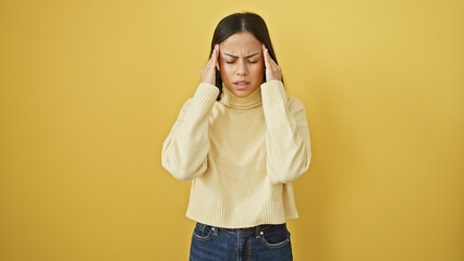 A distressed young hispanic woman in a cream sweater holding her head against a yellow background.