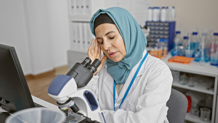 A middle-aged woman scientist in a lab coat using a microscope in a laboratory, looking tired or with a headache.