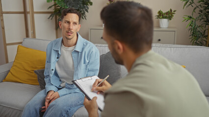 Man in a light blue shirt seated on a sofa discusses with another man taking notes in a cozy living room setting surrounded by plants and cushions.