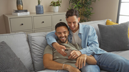 Two men embracing on a sofa at home, smiling and looking at a phone, depicting a loving gay couple in a cozy living room with plants and modern decor