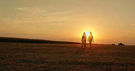 A young farming couple walks through a field at sunset, while a combine harvests crops in the...