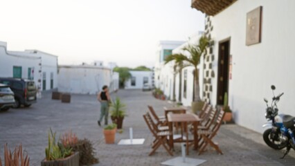 Blurred woman walking outdoors in lanzarote, spain with defocused background featuring white buildings, potted plants, parked cars, and outdoor seating on a paved street