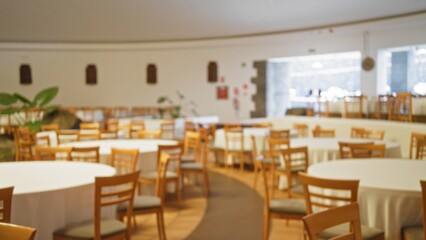 Blurred empty wedding hall with round tables and wooden chairs set for an event, creating a defocused bokeh effect in a spacious restaurant setting.