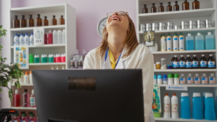 A young, attractive blonde woman in a white coat laughs joyfully while working in a well-stocked pharmacy.