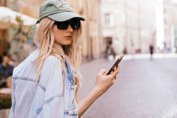 Beautiful smiling woman walking on street chatting on mobile phone. Girl talking on smartphone, texting, sending message, green cap, sun glasses and jeans jacket. Girl turn her head.