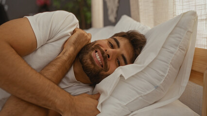 Young hispanic man with a beard lying on a bed in a cozy bedroom, smiling and looking relaxed in a comfortable home setting.