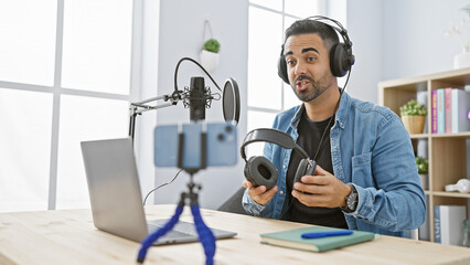 Handsome hispanic man with beard hosting podcast in modern studio wearing headset and holding...