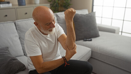 Fototapeta premium Grey-haired man with a beard sitting in a living room examining his arm, in a modern apartment interior with a light grey couch and a plant in the background.