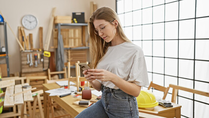 A blonde woman examines her smartphone in a well-lit carpentry workshop full of tools and wooden materials.