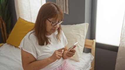 An elderly hispanic woman sits on a bed in her bedroom, using a smartphone, dressed in casual white clothes and pink pajamas, with natural light streaming into the cozy room.