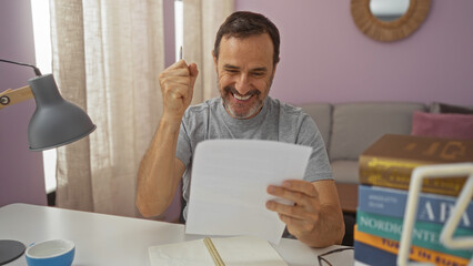 Happy mature man celebrating success at home while reading documents in a cozy living room with a stack of books and a cup of coffee on the table.