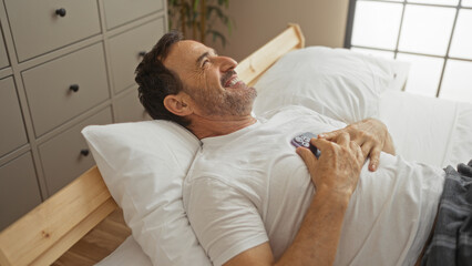 Obraz premium Handsome hispanic man lying in a bed with a cell phone held to his chest, smiling in a cozy bedroom setting.