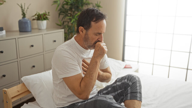 Middle-aged hispanic man sitting on bed in bedroom holding his chest while coughing, surrounded by indoor plants and furniture. - Powered by Adobe