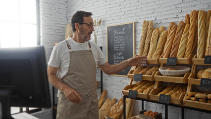 Mature hispanic man in a bakery arranging bread on wooden shelves, dressed in an apron, in a bright...