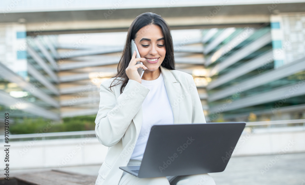 Poster A woman engaged in a phone call while using her laptop, smiling in an outdoor workspace surrounded by modern architecture.