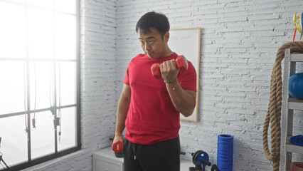 A handsome young chinese man in a gymnasium focuses on lifting red dumbbells, showcasing indoor...