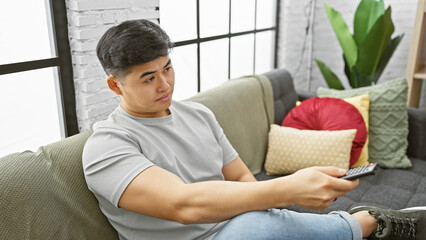 A young asian man lounges on a couch in a modern apartment, holding a remote control with a pensive expression.