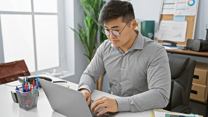 A young asian man working attentively on his laptop in a modern office setting.