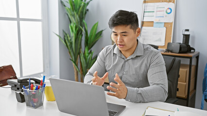 Young asian man working on laptop in modern office, expressing problem-solving determination.