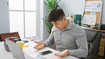 A perplexed young asian man in a modern office setting looking at a smartphone with a laptop and coffee on the desk.