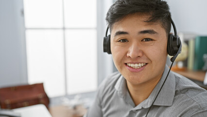A young asian man with a headset smiling in an office setting, suggesting a customer service or helpdesk representative.