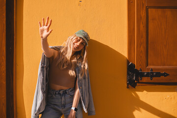 Attractive close up portrait of pretty blonde woman covering her face on yellow wall. Happy girl hiding from the sun light at sunny day with five fingers hand. Smiling girl wear cap, jeans jacket.