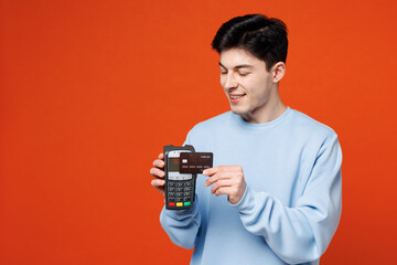 Young fun man he wears blue sweatshirt casual clothes hold wireless modern bank payment terminal to process acquire credit card isolated on plain red orange color background studio. Lifestyle concept.