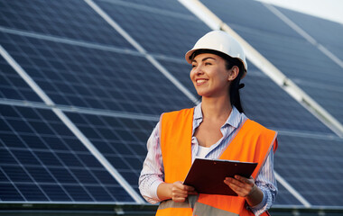 Notepad in hands. Female worker engineer in uniform is near solar panels - Powered by Adobe