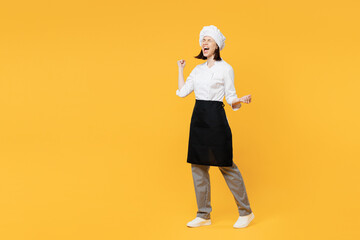 Full body young chef cook baker woman wear white shirt black apron uniform toque chefs hat doing winner gesture celebrate clench fists say yes isolated on plain yellow background Cooking food concept