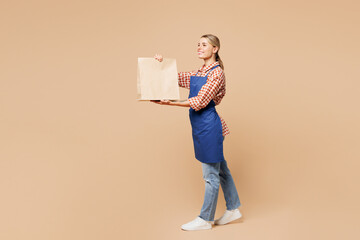 Full body young woman barista bartender barman employee wear blue apron red shirt work in coffee shop hold craft paper takeaway bag isolated on plain beige background. Small business startup concept.